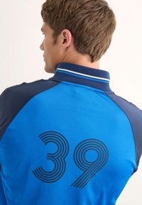 Blå pikétröja med marinblå detaljer, med ett strukturerat nummer "39" i böljande linjer. Kragdesign med ribbad halslinning.