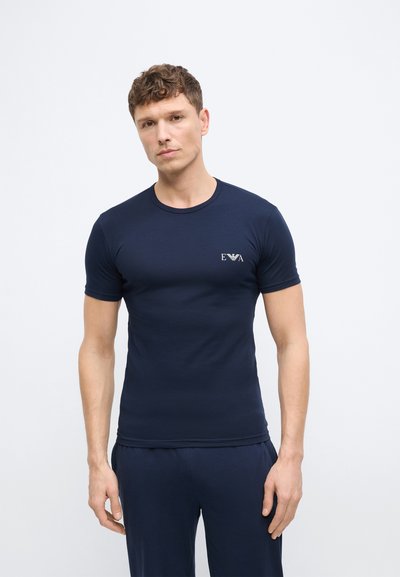 Άνδρας που φοράει εφαρμοστό ναυτικό μπλε μπλουζάκι με το λογότυπο "EVA" στο στήθος, στέκεται μπροστά σε λευκό φόντο, κοιτάζοντας μπροστά με ουδέτερη έκφραση.