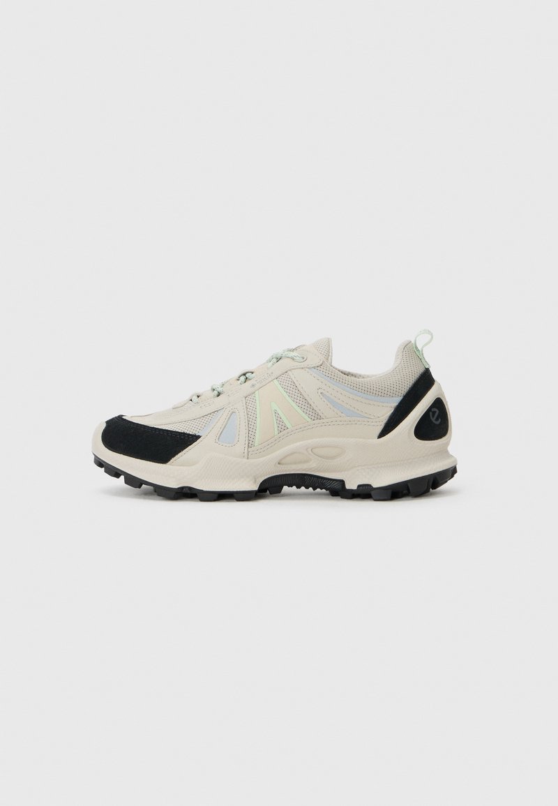 Helle beige Sneaker mit schwarzen Wildlederakzenten, Obermaterial aus Mesh, reflektierenden Details und strukturiertem Gummisohle für besseren Halt. Schnürdesign.