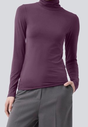 Femme portant un haut violet ajusté à col roulé à manches longues et un pantalon gris, une main dans la poche et l'autre reposant sur le côté.