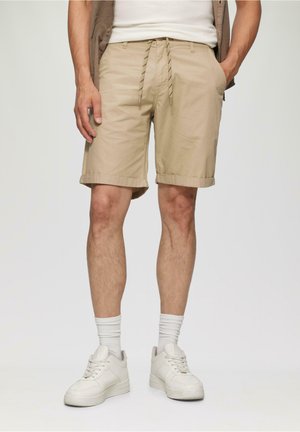 Shorts beiges mi-mollet avec ourlets retroussés, taille élastique à cordon de serrage et poches latérales, associés à des baskets blanches et des chaussettes côtelées.