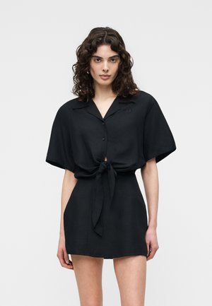 Göndör hajú nő fekete, rövid ujjú, gombos felsőben, amelyet a deréknál megkötött, és hozzá illő, magasderekú szoknyát visel.