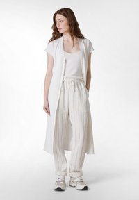 Langer weißer Weste über einem weißen Tanktop, kombiniert mit hell gestreiften Hosen und beigen Sneakers. Weicher Stoff, lässige Passform und Knopfdetails.