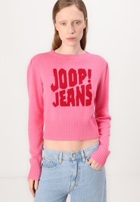 Pink cropped sweater with ribbed hem and cuffs. Features bold red knitted text "JOOP! JEANS" on the front. Soft texture, casual design.