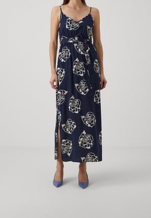 Kobieta w granatowej długiej sukience w białe kwiatowe wzory, z cienkimi ramiączkami, rozcięciem po boku, przewiązana paskiem w talii, w niebieskich szpiczastych szpilkach.