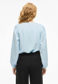 Blouse bleu clair à manches bouffantes, texture lisse et large encolure, associée à un pantalon noir avec taille élastique et poches arrière.