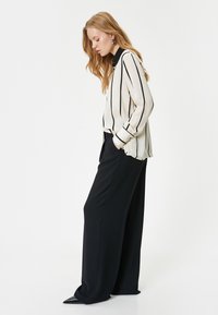 Striped blouse in cream and black, features long sleeves and a collar. Paired with high-waisted, wide-leg black trousers and pointed shoes.