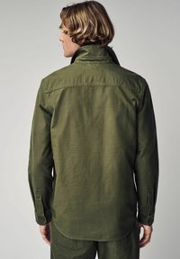 Olive green long-sleeve shirt with a collar, buttoned cuffs, and a straight hem. Features a smooth, textured fabric with minimal design details.