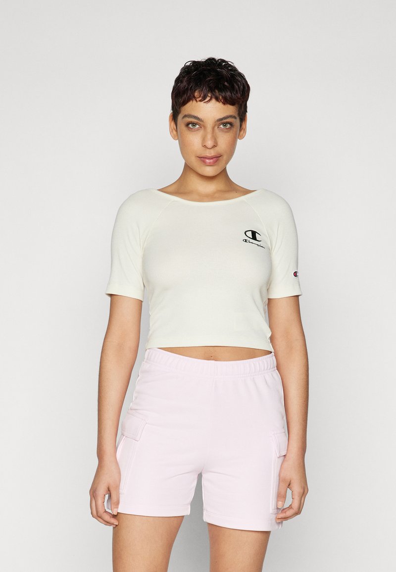 Lys beige, kortermet t-skjorte med ribbestruktur; har en liten svart logo. Kombinert med lys rosa shorts med elastisk midje.