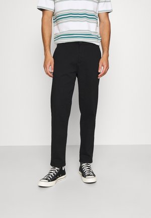 Homme portant un pantalon noir, des baskets montantes noires et une chemise à manches courtes blanche avec des rayures horizontales vertes et grises.