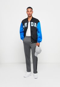 Sort og blå varsity jakke med "JORDAN" bogstaver, hvid t-shirt, grå bukser og grå baseballcap; atletiske sko fuldender outfittet.