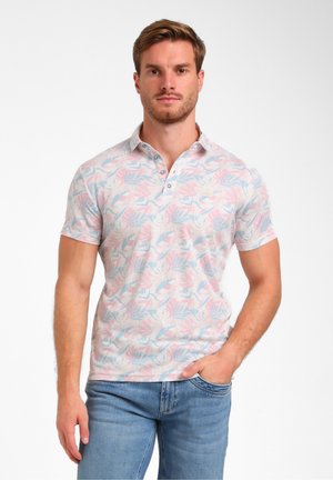 Giovane uomo con capelli castano chiaro che indossa una polo a fiori rosa pastello e blu e jeans blu, in piedi su sfondo bianco.