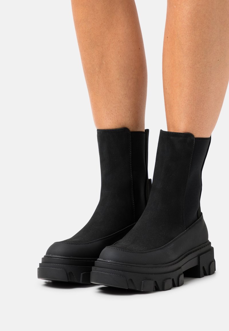 Bottes noires à plateforme épaisse montant jusqu'au milieu du mollet, avec semelles crantées épaisses, portées sur des jambes nues contre un fond clair uni.