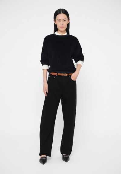 Μαύρο φούτερ με λευκά μανσέτα, σε συνδυασμό με μαύρο παντελόνι με φαρδιά γραμμή και καφέ ζώνη. Τα παπούτσια είναι μυτερά μαύρα με γυαλιστερό φινίρισμα.