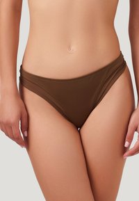 Bas de bikini marron avec une texture lisse, présentant un design échancré et des fronces sur les côtés pour plus de détails. Fabriqué en matériau de maillot de bain extensible.