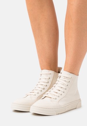 Piernas con zapatillas de lona de caña alta color blanco sucio con cordones, de pie sobre una superficie blanca contra un fondo liso.