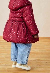 Chaqueta de plumas roja con un patrón de estrellas, cintura elástica y cierre de cremallera, combinada con unos vaqueros de mezclilla azules y zapatillas doradas.