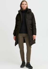 Padded black parka jacket with a hood, featuring a zip closure and front pockets. Paired with a knitted black sweater and green pants.