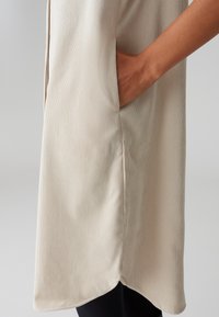 Tunic in tessuto beige a coste con tasca laterale e orlo arrotondato, caratterizzato da una superficie leggermente testurizzata.