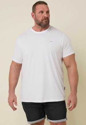 Bärtiger Mann mit kurzem Haar, der ein schlichtes weißes T-Shirt und schwarze hochgekrempelte Shorts trägt, steht und schaut nach links vor neutralem Hintergrund.