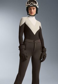 Lyžiarsky oblek s vysokým golierom, ktorý má rebrovaný dizajn v čiernej, krémovej a šedej farbe. Nosí sa s hrubými čiernymi rukavicami a lyžiarskou prilbou.