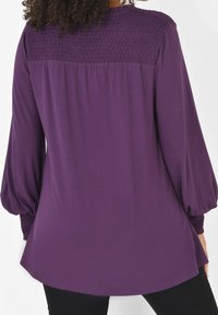 Violetti pitkähihaisen paita, jossa on rypytetty yksityiskohta olkapäillä, kaareva helma ja sileä pinta. Hihoissa on puffeja ja ne on kerätty ranteiden ympärille.