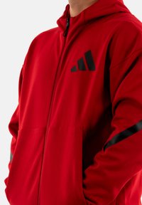 Felpa rossa con zip realizzata in tessuto morbido, dotata di un logo nero e dettagli sulle maniche. Include un cappuccio e tasche frontali.