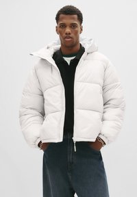Jonge man in een witte gewatteerde jas over een zwart shirt en blauwe jeans, staand met zijn handen in zijn zakken tegen een effen achtergrond.