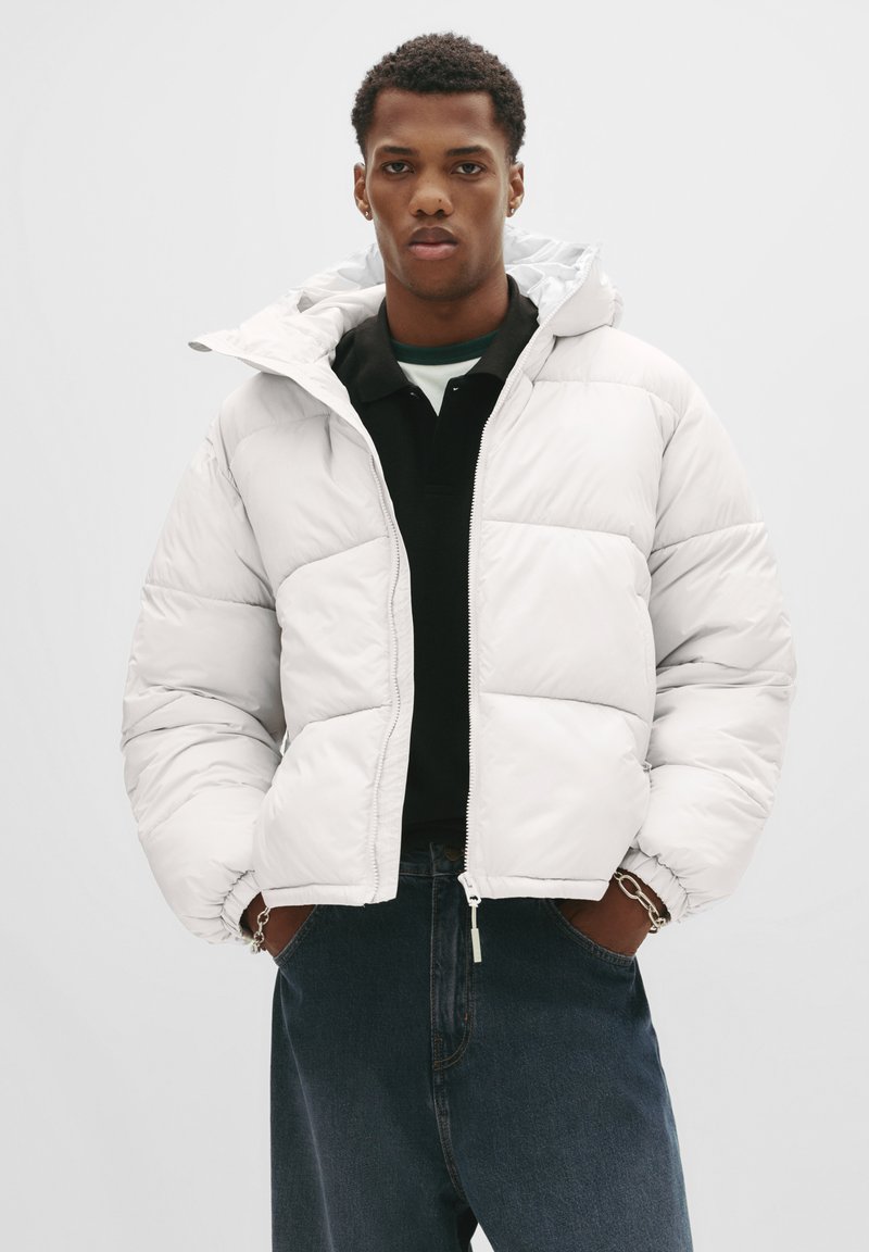 Jonge man in een witte gewatteerde jas over een zwart shirt en blauwe jeans, staand met zijn handen in zijn zakken tegen een effen achtergrond.