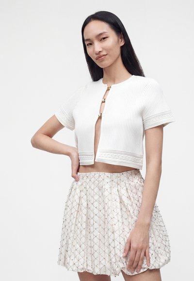 Jeune femme aux longs cheveux noirs porte un crop top blanc à manches courtes avec des fermoirs dorés et une jupe à taille haute et motifs.