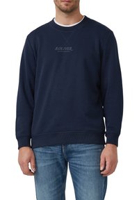 Uomo che indossa un sudore a maniche lunghe di colore blu navy con il logo e il testo "S.Oliver", abbinato a jeans blu di lavaggio medio, in piedi su uno sfondo bianco.