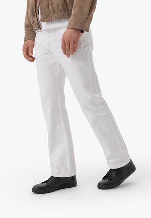 Uomo che indossa pantaloni bianchi, sneakers nere e una giacca marrone, in piedi su uno sfondo bianco uniforme.
