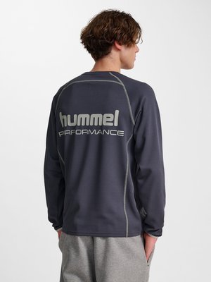 Ung mand med krøllet hår iført en mørkegrå langærmet top med teksten "hummel PERFORMANCE" på ryggen og lysegrå bukser.