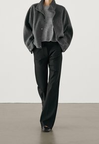 Veste en laine grise avec des boutons pression, associée à un pull gris et un pantalon large noir en tissu lisse. Chaussures noires à enfiler.