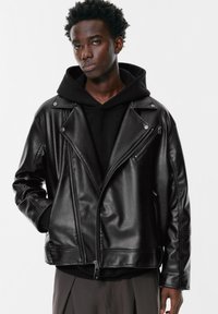 Crna kožna bikerska jakna s asimetričnim patentnim zatvaračem, kopčama na dugmad i opuštenim krojem, složena preko crnog dukserice.