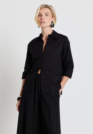 Blonde Frau trägt schwarzes Lochmuster-Hemd mit 3/4-Ärmeln und schwarze, weit geschnittene Hosen, posiert mit einer Hand in der Tasche.