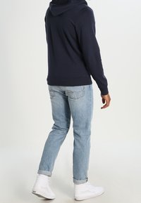 Felpa navy con grande cappuccio, jeans azzurri chiari e sneaker alte bianche. Cuciture visibili sui jeans e sulle sneaker. Look casual.