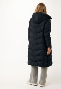 Černá péřová bunda s kapucí, s chevronovým prošíváním, dlouhým střihem a měkkou texturou. V kombinaci se světle šedými flared kalhotami a teniskami.