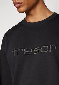 Felpa nera con collo rotondo, caratterizzata da un logo "tresor" ricamato e testurizzato in grigio scuro sulla parte anteriore.