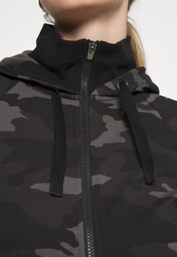 Haut de sweat à capuche zippé en camouflage noir et gris avec un col haut, une texture contrastante et des cordons ajustables à la capuche.