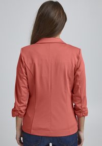 Blazer corail au fit ajusté, épaules structurées et manches retroussées. Tissu lisse avec une texture douce, comportant une couture centrale.