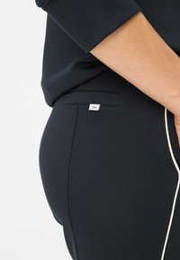 Zwarte stretchbroek met een slanke pasvorm, voorzien van een witte accentstreep en een klein merkje op de tailleband. Gladde textuur.