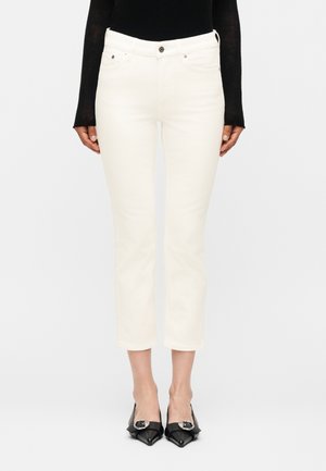 Γυναίκα με λευκά κοντά τζιν, μαύρο μακρυμάνικο τοπ και μαύρες μυτερές γόβες με ασημένιες αγκράφες, στέκεται μπροστά σε απλό φόντο.