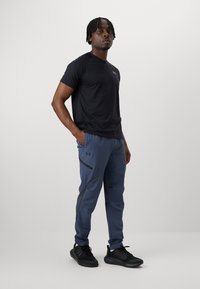 T-shirt de sport noir à manches courtes avec un logo, associé à un pantalon de jogging bleu fuselé avec une poche zippée et des accents noirs. Des baskets noires complètent la tenue.