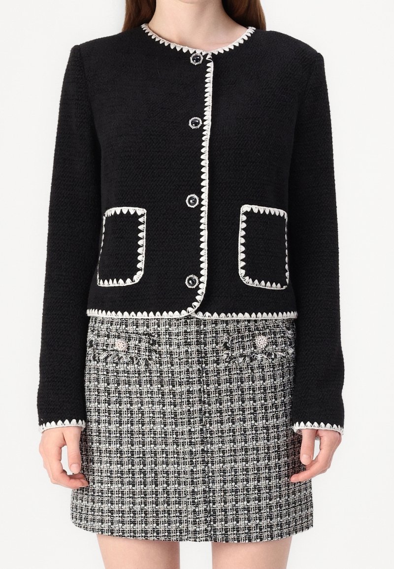 Giacca nera strutturata con rifiniture e bottoni a zigzag bianchi, abbinata a una gonna in tweed bianca e nera con tasche decorativa.