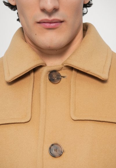 Μπεζ παλτό από μάλλινο μείγμα με φαρδιά γιακά, δύο κουμπιά και απαλή υφή. Τα κουμπιά είναι σκούρο καφέ με διακριτική λάμψη.