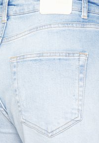 Jeans de denim azul claro com um único bolso traseiro. As características incluem um rótulo branco da marca na cintura e detalhes de costura subtis.