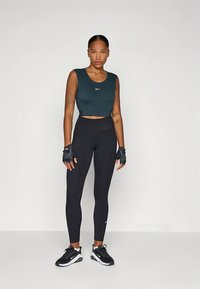 Tmavě zelený crop top s bílým logem, černé legíny s vysokým pasem, rukavice na cvičení bez prstů a černé sportovní boty.