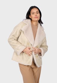 Cappotto beige doppiopetto con colletto e polsini in pelliccia sintetica, texture morbida e due grandi bottoni frontali; indossato sopra un maglione a coste.