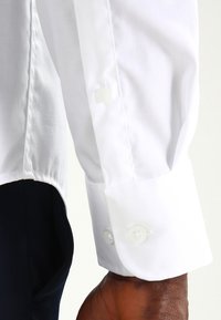 Camisa blanca de manga larga con puños abotonados, que presenta una textura suave y un ajuste entallado, con una tela oscura parcialmente visible debajo.